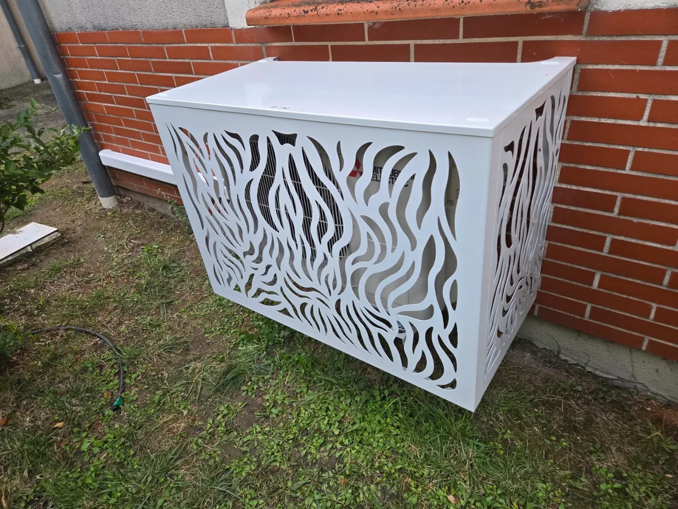 Cache décoratif blanc autour d’un groupe de climatisation devant une façade en briques